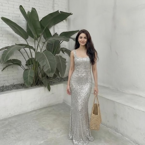 May include: A silver sequin gown with a square neckline and a fitted silhouette. The dress is floor-length and shimmers. A woven tote bag is held in the hand. The background features a large green plant and a white wall.