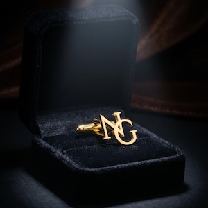 May include: A gold-toned metal cufflink featuring the interlocking capital letters N and G. The cufflink is displayed inside a black velvet-lined jewelry box, which is open to reveal the metallic finish and the monogram design clearly.
