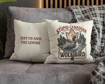 May include: Two off-white throw pillows with text and graphics. One pillow reads "JUST US AND THE LOONS" in brown text. The other pillow features a graphic of a loon in a lake with the text "STUPID CANADIAN WOLF BIRD".