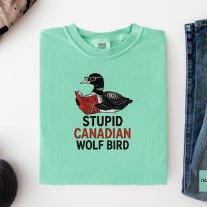 May include: A light green t-shirt with a graphic of a bird wearing glasses and reading a book. The text on the shirt reads "STUPID CANADIAN WOLF BIRD" in black and red. The shirt is next to a pair of blue jeans.