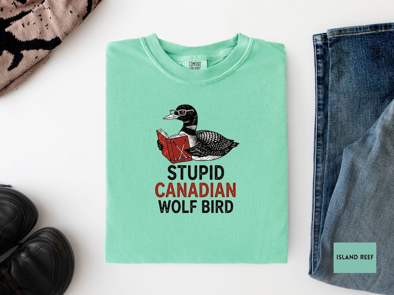 May include: A light green t-shirt with a graphic of a bird wearing glasses and reading a book. The text on the shirt reads "STUPID CANADIAN WOLF BIRD" in black and red. The shirt is next to a pair of blue jeans.