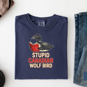 May include: Navy blue t-shirt with a graphic of a loon wearing glasses and reading a book. The text "STUPID CANADIAN WOLF BIRD" is printed below the graphic in red and white. The shirt is folded on a white surface.