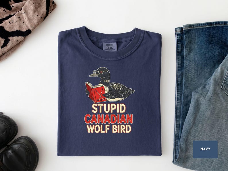 May include: Navy blue t-shirt with a graphic of a loon wearing glasses and reading a book. The text "STUPID CANADIAN WOLF BIRD" is printed below the graphic in red and white. The shirt is folded on a white surface.
