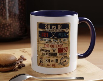 May include: A white ceramic mug with a blue interior and handle. The mug features a vintage-style ticket design with text including "SH vs IR," game details, and "The Cottage." A spoon and a pastry are in the foreground.