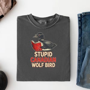 May include: Dark gray t-shirt with a graphic of a loon wearing glasses and reading a book. The text "STUPID CANADIAN WOLF BIRD" is printed below the graphic in red and white. The shirt is laid flat on a white surface.