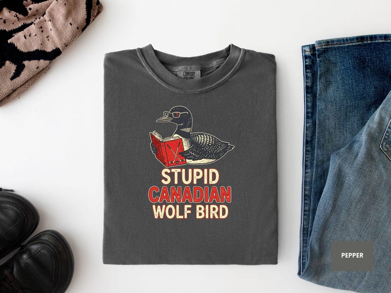 May include: Dark gray t-shirt with a graphic of a loon wearing glasses and reading a book. The text "STUPID CANADIAN WOLF BIRD" is printed below the graphic in red and white. The shirt is laid flat on a white surface.