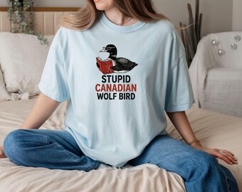 May include: Light blue t-shirt featuring a cartoon illustration of a black and white bird wearing glasses and reading a book. The text "STUPID CANADIAN WOLF BIRD" is printed below the illustration. The shirt is made of a soft material.