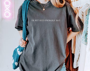 May include: Dark gray t-shirt with the text "OK. NOT GOOD. PROBABLY BAD." in white. The shirt is a casual style, with short sleeves. A blue knitted bag with white heart patterns is visible.