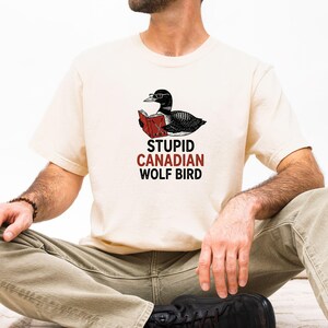 May include: Cream-colored t-shirt featuring a black and white illustration of a loon wearing glasses and reading a book. The text "STUPID CANADIAN WOLF BIRD" is printed below the bird in red and black.