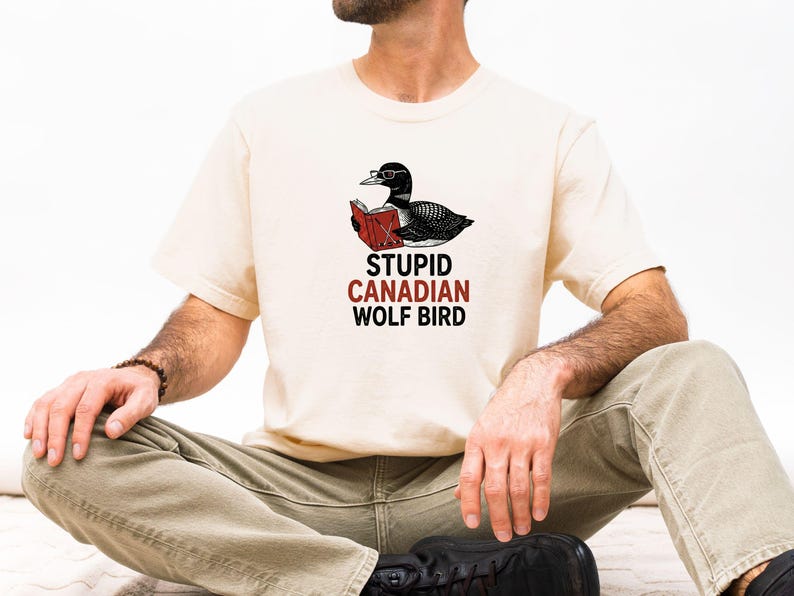 May include: Cream-colored t-shirt featuring a black and white illustration of a loon wearing glasses and reading a book. The text "STUPID CANADIAN WOLF BIRD" is printed below the bird in red and black.