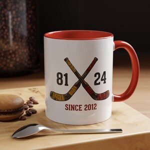 May include: White ceramic mug with a red interior and handle. The mug features crossed hockey sticks with the numbers 81 and 24, and the text "SINCE 2012" in red. A spoon and a pastry are in the foreground.