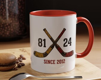 May include: White ceramic mug with a red interior and handle. The mug features crossed hockey sticks with the numbers 81 and 24, and the text "SINCE 2012" in red. A spoon and a pastry are in the foreground.