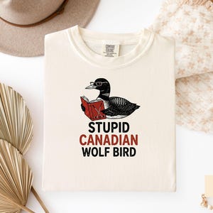 May include: Ivory-colored t-shirt featuring a cartoon loon wearing glasses and reading a book. The text "STUPID CANADIAN WOLF BIRD" is printed below the illustration in black and red. A hat and decorative fan are in the background.