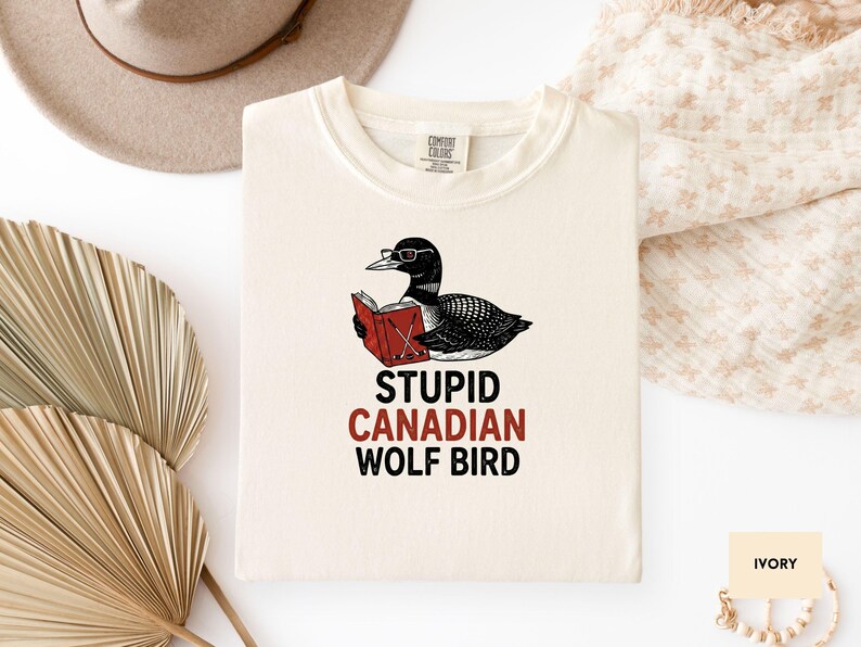 May include: Ivory-colored t-shirt featuring a cartoon loon wearing glasses and reading a book. The text "STUPID CANADIAN WOLF BIRD" is printed below the illustration in black and red. A hat and decorative fan are in the background.
