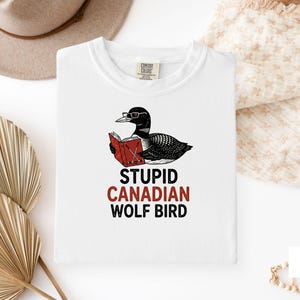May include: White t-shirt featuring a black and white illustration of a bird wearing glasses and reading a red book. The text "STUPID CANADIAN WOLF BIRD" is printed below the illustration in black and red.