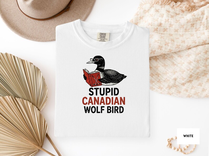 May include: White t-shirt featuring a black and white illustration of a bird wearing glasses and reading a red book. The text "STUPID CANADIAN WOLF BIRD" is printed below the illustration in black and red.