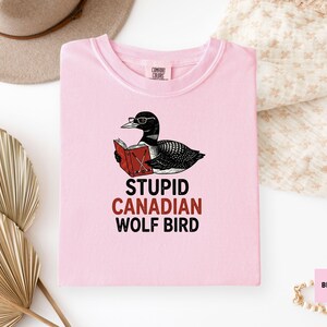 May include: A light pink t-shirt featuring a cartoon illustration of a loon wearing glasses and reading a book. The text "STUPID CANADIAN WOLF BIRD" is printed below the illustration. The shirt is made of a soft material.