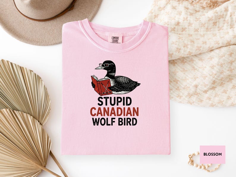May include: A light pink t-shirt featuring a cartoon illustration of a loon wearing glasses and reading a book. The text "STUPID CANADIAN WOLF BIRD" is printed below the illustration. The shirt is made of a soft material.