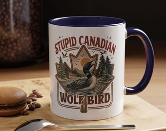 May include: White ceramic mug with a blue interior and handle. The mug features a graphic of a loon in a lake, surrounded by trees and the text "STUPID CANADIAN WOLF BIRD". A spoon and a pastry are in the foreground.