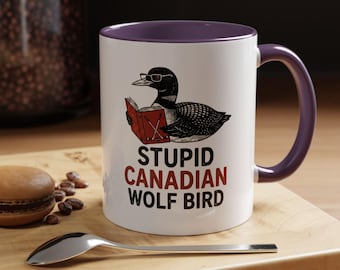 May include: White ceramic mug with a purple interior and handle. The mug features a cartoon illustration of a loon wearing glasses and reading a book. Text on the mug reads "STUPID CANADIAN WOLF BIRD".