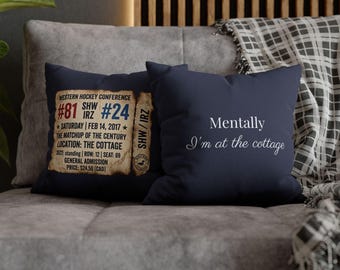 May include: Two navy blue throw pillows on a gray couch. One pillow features text from the Western Hockey Conference, including the date, teams, and location. The other pillow has the text "Mentally I'm at the cottage."