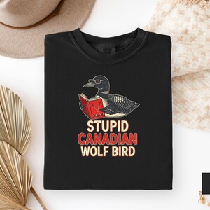 May include: Black crew neck t-shirt featuring a cartoon loon wearing glasses and reading a book. The text "STUPID CANADIAN WOLF BIRD" is printed below the illustration in red and white. The shirt is displayed on a white surface.