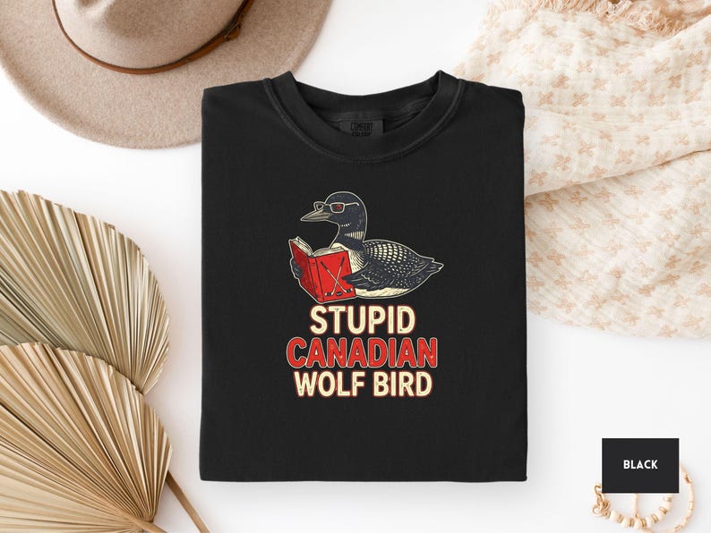 May include: Black crew neck t-shirt featuring a cartoon loon wearing glasses and reading a book. The text "STUPID CANADIAN WOLF BIRD" is printed below the illustration in red and white. The shirt is displayed on a white surface.