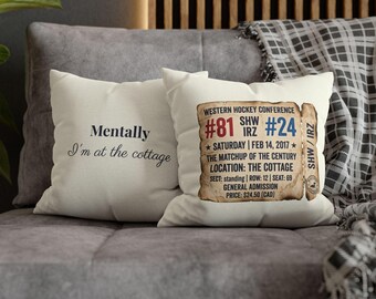 May include: Two off-white throw pillows on a gray couch. One pillow has the text "Mentally I'm at the cottage" in a script font. The other pillow features a vintage-style ticket with text about a hockey conference.