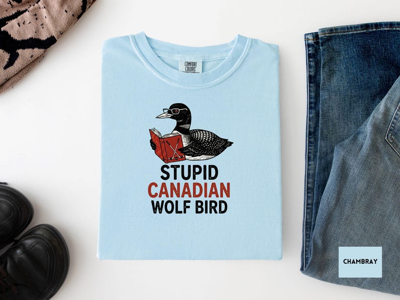 May include: Light blue t-shirt with a graphic of a loon wearing glasses and reading a red book. The text "STUPID CANADIAN WOLF BIRD" is printed below the graphic. The shirt is folded and placed on a white surface with jeans and shoes.