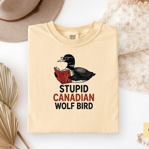 May include: Cream-colored t-shirt featuring a cartoon loon wearing glasses and reading a book. The text "STUPID CANADIAN WOLF BIRD" is printed below the graphic in black and red. The shirt is folded on a white surface.