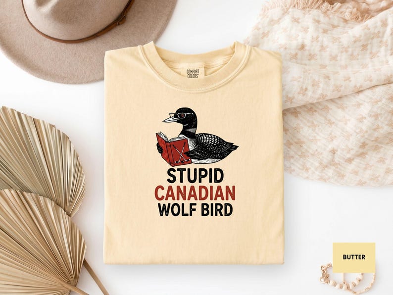 May include: Cream-colored t-shirt featuring a cartoon loon wearing glasses and reading a book. The text "STUPID CANADIAN WOLF BIRD" is printed below the graphic in black and red. The shirt is folded on a white surface.