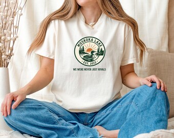 May include: Cream-colored t-shirt featuring a circular graphic with the text "Muskoka Lake EST. 2012" and a nature scene with a duck. The shirt also has the phrase "WE WERE NEVER JUST RIVALS" below the graphic. The model is wearing blue jeans.