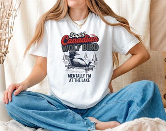 May include: White t-shirt with the text "Stupid Canadian Wolf Bird" in red, black, and white. The graphic shows a cartoon bird relaxing on a beach chair. The text "Mentally I'm at the lake" is below the graphic.