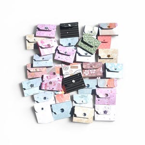 May include: A collection of small, colorful paper pouches with various patterns, including floral, striped, and geometric designs. Each pouch is secured with a button closure. The pouches are in shades of pink, blue, green, and white.