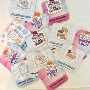 May include: A collection of magnetic bookmarks with various designs and text. Some bookmarks feature illustrations of a book with flowers, a clipboard, and a bear with coffee. Other bookmarks have the phrases "Pray more, worry less" and "Dogs make our lives complete."