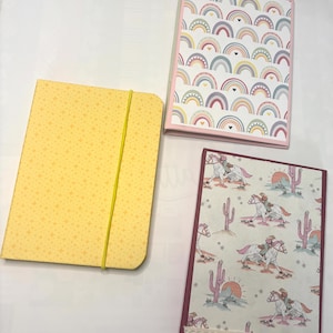 Può includere: Tre quaderni con copertine diverse. Uno è giallo con un motivo a stelle e un elastico giallo. Un altro ha un motivo arcobaleno su un bordo rosa. Il terzo ha un motivo di cowboy e cavallo con un bordo in pizzo.