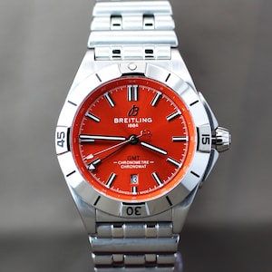 Könnte beinhalten: Eine Breitling Chronomat GMT Armbanduhr aus Edelstahl mit einem leuchtend orangefarbenen Zifferblatt. Die Uhr verfügt über ein silberfarbenes Metallgliederarmband, eine drehbare Lünette mit eingravierten Ziffern, leuchtende Zeiger, ein Datumsfenster auf der Sechs-Uhr-Position sowie den Schriftzug Breitling 1884, GMT, Chronometre und Chronomat.