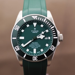 May include: A Tudor Pelagos watch with a green dial and matching green rubber strap. The watch features a black rotating bezel with white numerals, a date window at the 3 o'clock position, and white luminous markers. The dial text reads Tudor Geneve Pelagos Chronometer Officially Certified Rotor Self-Winding 500m-1640ft Swiss Made.