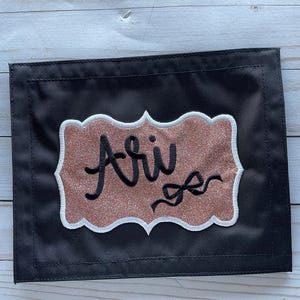 May include: A black fabric name tag with a white border and a pink glitter background. The name "Ari" is embroidered in black cursive with a black bow.