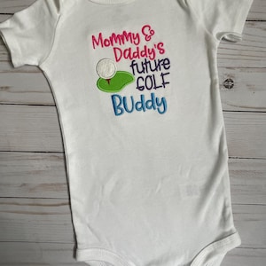 May include: White baby onesie with embroidered text that reads "Mommy & Daddy's future golf buddy" with a golf ball and green.