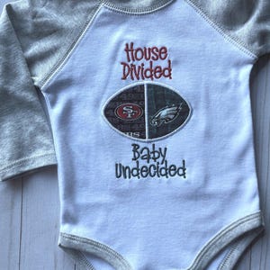 May include: White and gray baby onesie with raglan sleeves. The onesie features the text "House Divided" in red and "Baby Undecided" in green. An embroidered football graphic displays the logos of the San Francisco 49ers and the Philadelphia Eagles.