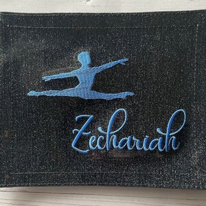 May include: A black fabric tote bag with a blue embroidered silhouette of a dancer and the name "Zechariah" in blue cursive lettering.