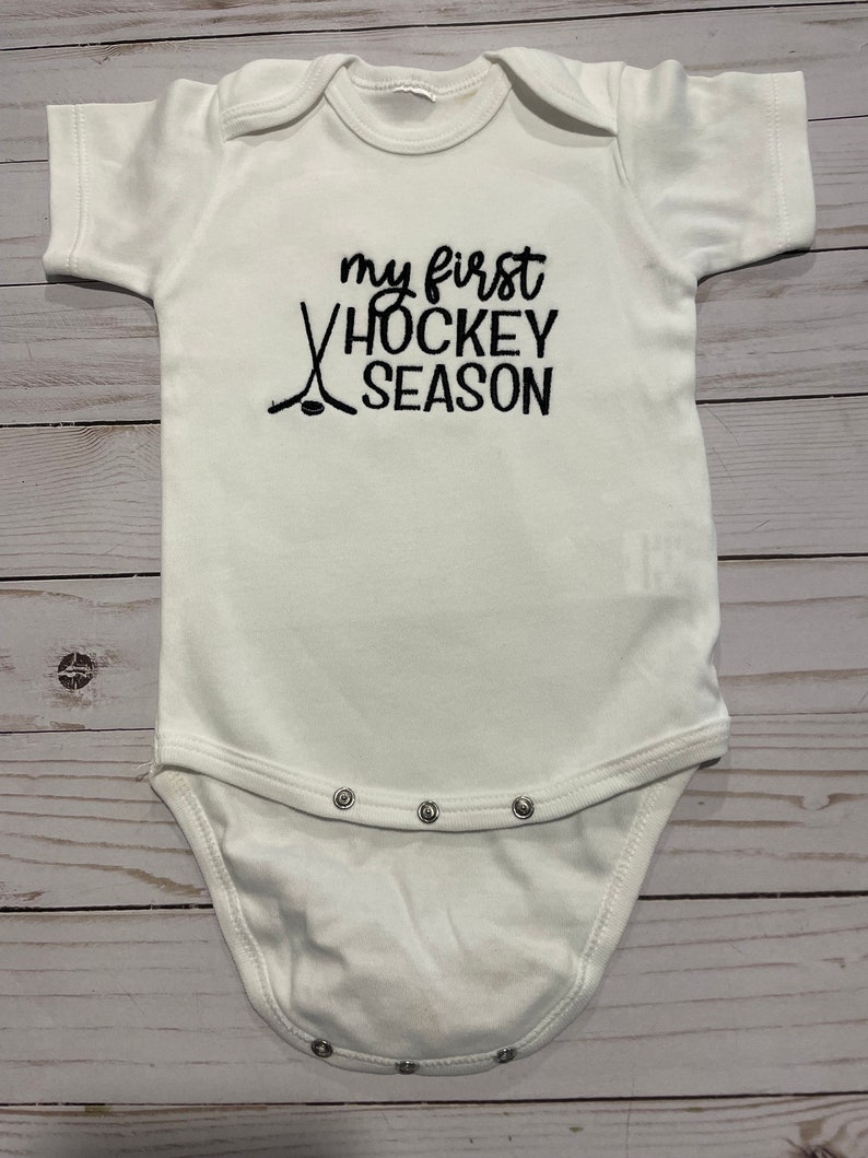 Puede incluir: Un body blanco para beb&eacute; con texto bordado en negro que dice "my first hockey season" con un par de palos de hockey y un disco.