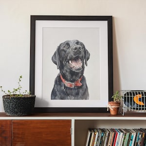 May include: A framed watercolor painting of a black Labrador Retriever with a red collar. The dog is smiling with its mouth open. The painting is in a black frame and is displayed on a wooden shelf.