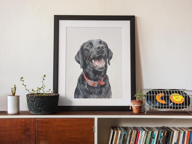 May include: A framed watercolor painting of a black Labrador Retriever with a red collar. The dog is smiling with its mouth open. The painting is in a black frame and is displayed on a wooden shelf.