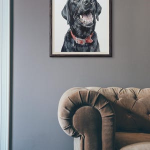 May include: A framed watercolor painting of a black Labrador Retriever with a red collar. The dog is smiling with its mouth open. The painting is in a dark wooden frame, hanging on a gray wall. A brown sofa is in the foreground.