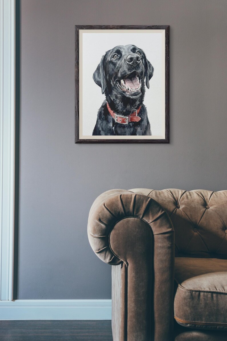 May include: A framed watercolor painting of a black Labrador Retriever with a red collar. The dog is smiling with its mouth open. The painting is in a dark wooden frame, hanging on a gray wall. A brown sofa is in the foreground.