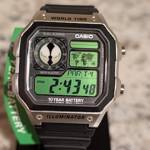Op de afbeelding: Een digitaal Casio-horloge met een zwarte kunststof band en een zilverkleurige kast. De wijzerplaat heeft een groen digitaal display met een wereldkaart, de tekst WORLD TIME, 5 ALARMS, WR100M, 10 YEAR BATTERY en ILLUMINATOR. Een rond grafisch icoon is zichtbaar.