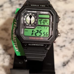 Może przedstawiać: Czarny zegarek cyfrowy Casio z zielonkawym wyświetlaczem, zawierający grafikę mapy świata, okrągłą tarczę pomocniczą w stylu analogowym z biało-czarnym logo oraz napis World Time, 5 Alarms, WR100M, 10 Year Battery i Illuminator na tarczy.