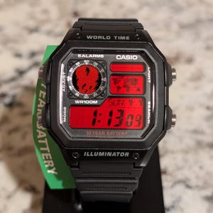 May include: A black Casio digital watch with a red display screen featuring a world map, analog-style clock face, and digital time readout. The watch face includes text reading WORLD TIME, 5 ALARMS, CASIO, WR100M, 10 YEAR BATTERY, and ILLUMINATOR.